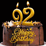 92nd birthday GIF featuring a chocolate birthday cake displaying 'Happy Birthday' lettering in gold, and two lit gold candles shaped as '92' lighting up the top.