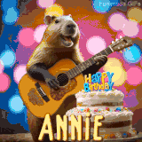 Animated birthday GIF showing a capybara playing guitar and singing beside a birthday cake with the name Annie in large letters and colorful bokeh lights in the background.