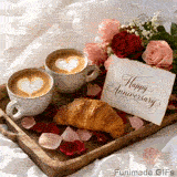 Two cappuccino cups with heart latte art sit on a wooden tray with a golden croissant, scattered rose petals, and a bouquet of red and pink roses on white sheets.