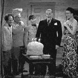 A black-and-white comedy scene where a birthday cake unexpectedly bursts, covering the man in cake while others laugh and react in surprise.