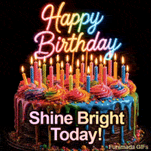 A chocolate birthday cake with vibrant rainbow drips, colorful frosting swirls, and many burning candles stands under a glowing neon Happy Birthday sign.