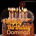 A rich chocolate cake topped with glowing birthday candles and surrounded by bright fireworks. The animated GIF displays the message 'Happy Birthday Domingo' in golden text.