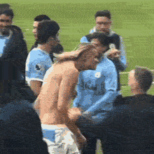A shirtless Erling Haaland with long blonde hair walks across a football pitch with a funny, intense, and hunched posture. Manchester City players stand in the background.