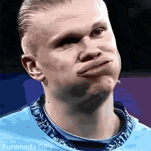 Close up of blonde footballer Erling Haaland puffing his cheeks out and pushing his lips forward in a funny fish face expression on a blurred blue background.