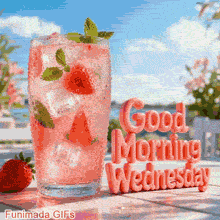 A refreshing summer drink with ice and strawberries sits on a bright outdoor table against a blue sky background. Glossy pink 3D letters beside the glass spell Good Morning Wednesday.