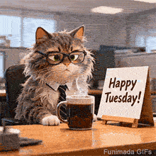 A fluffy tabby cat wearing black glasses and a professional blue tie sits at a wooden office desk next to a steaming mug of black coffee and a white sign that reads Happy Tuesday.